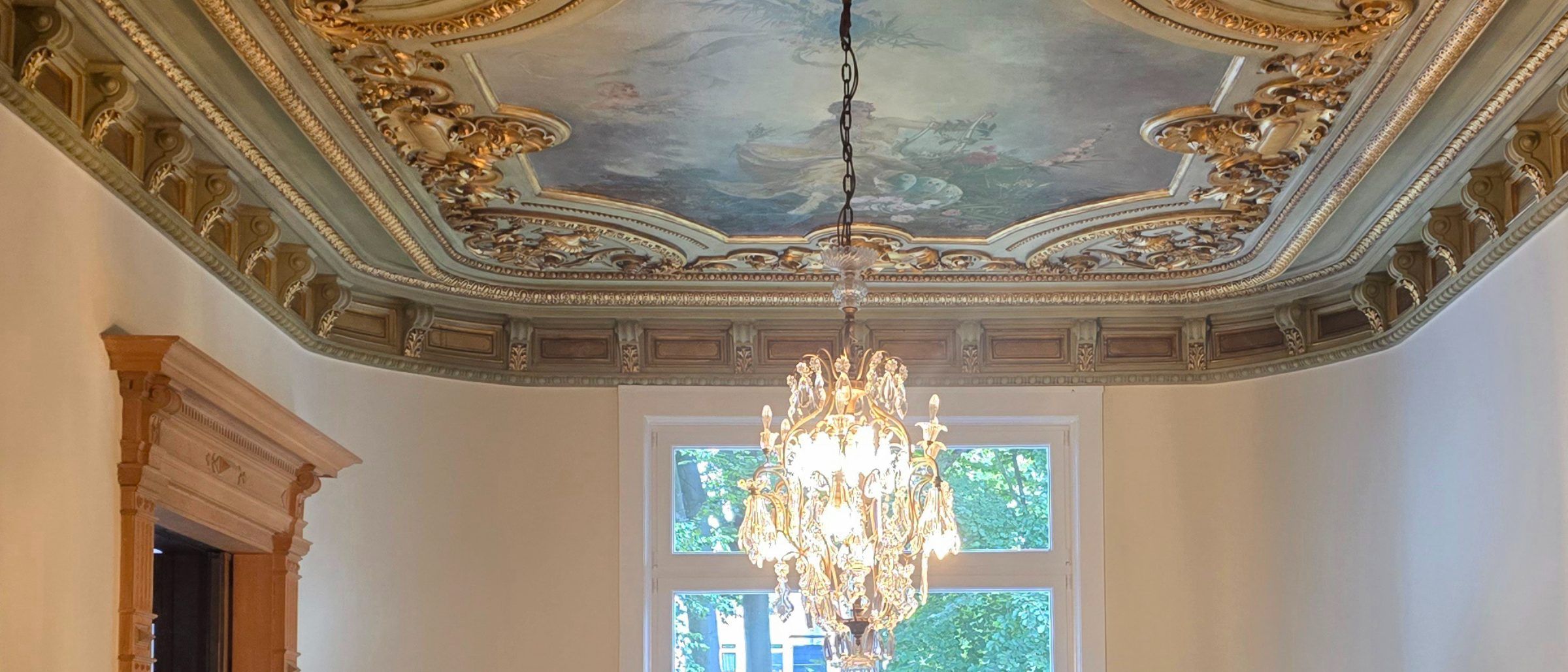 Der Saal der Villa: vergoldeter Stuck, Deckenmalerei, ein Kristalllüster vor dem hohen Fenster