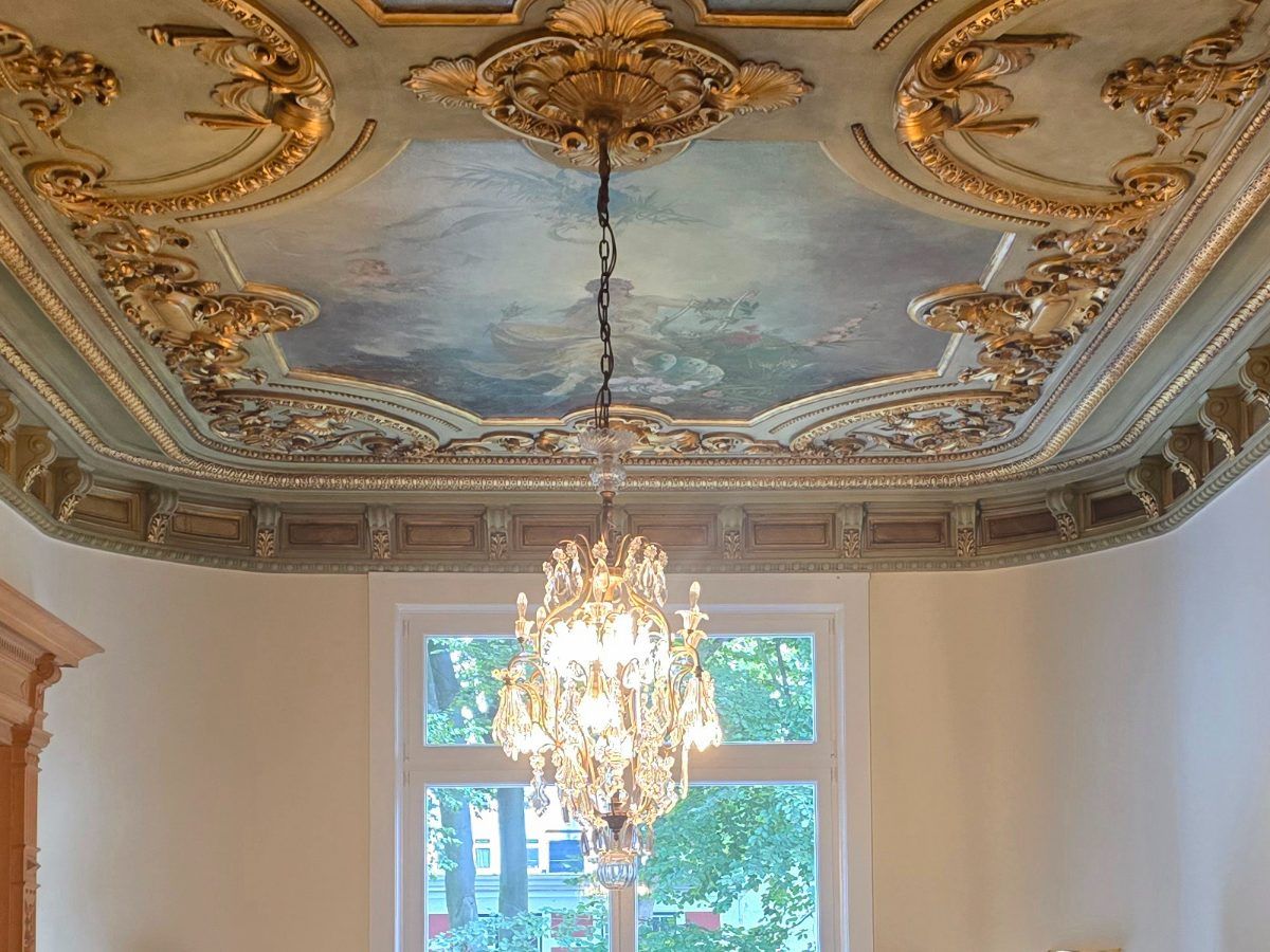 Der Saal der Villa: vergoldeter Stuck, Deckenmalerei, ein Kristalllüster vor dem hohen Fenster